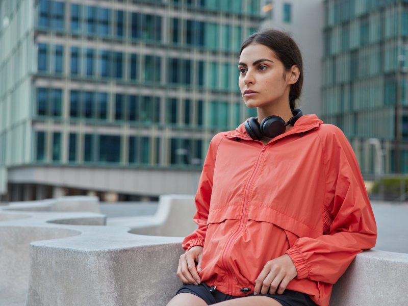 Young female in windbreaker leans with elbows on stone does exercises outdoors motivates to lead healthy lifestyle goes in for sport on city street looks forwards poses against urban building behind